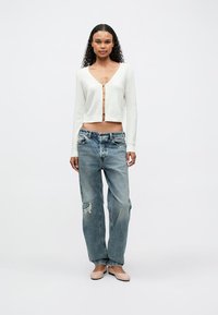 Cardigan blanc court avec un devant boutonné, associé à un jean bleu clair déchiré et des ballerines beiges. Le modèle se tient devant un fond uni.