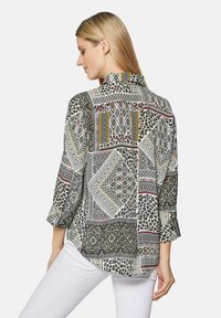 Musterbluse mit geometrischen Designs in Schwarz, Weiß und Rot, ergänzt durch einen Leopardenmusterakzent. Lange Ärmel mit Kragen und runder Saum.