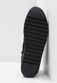 Semelle de chaussure en caoutchouc noir présentant une surface texturée avec des rainures horizontales et un logo circulaire. Le bord est doucement arrondi.