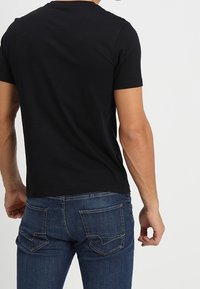 Hombre vestido con una camiseta negra de manga corta lisa y jeans ajustados azul oscuro, visto desde la parte de atrás contra un fondo blanco.