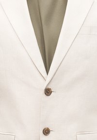 Μπεζ σακάκι με δύο καφέ κουμπιά και με πέτο με εγκοπή, φορεμένο πάνω από ένα ελιάς ρούχο από κάτω.