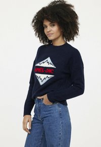 Herrenpullover in Marineblau mit weichem Textil, featuring ein weißes Bergdesign und roten "APRES-CHIC"-Text auf der Vorderseite. Kombiniert mit blauen hochgeschnittenen Jeans.