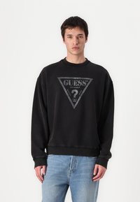 Melna kokvilnas sporta krekls ar apaļu dekoltē. Ierobežota pelēka trīsstūra logotipa apdruka ar vārdu "GUESS" centrā.