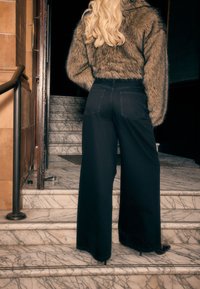 Sorte, vidde-ben denim bukser med høj talje, kombineret med en tekstureret brun imiteret pelsjakke, på en marmortrappe.