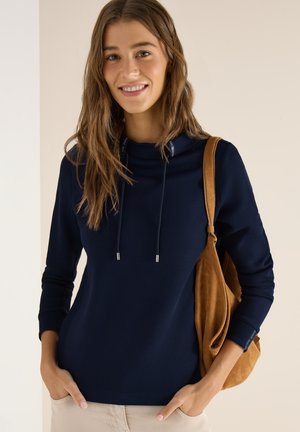 Giovane donna sorridente, indossa una felpa con cappuccio a maniche lunghe blu navy, pantaloni beige e porta una borsa a tracolla in camoscio marrone. Mani nelle tasche.