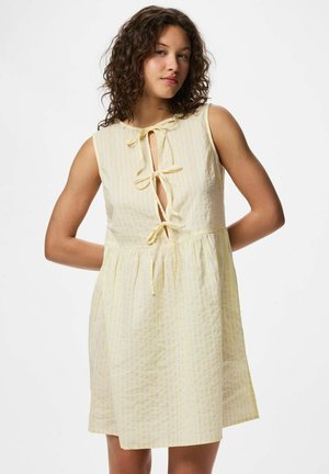 Jeune femme aux cheveux bouclés portant une robe sans manches jaune clair à rayures verticales avec des détails à nouer sur le devant.