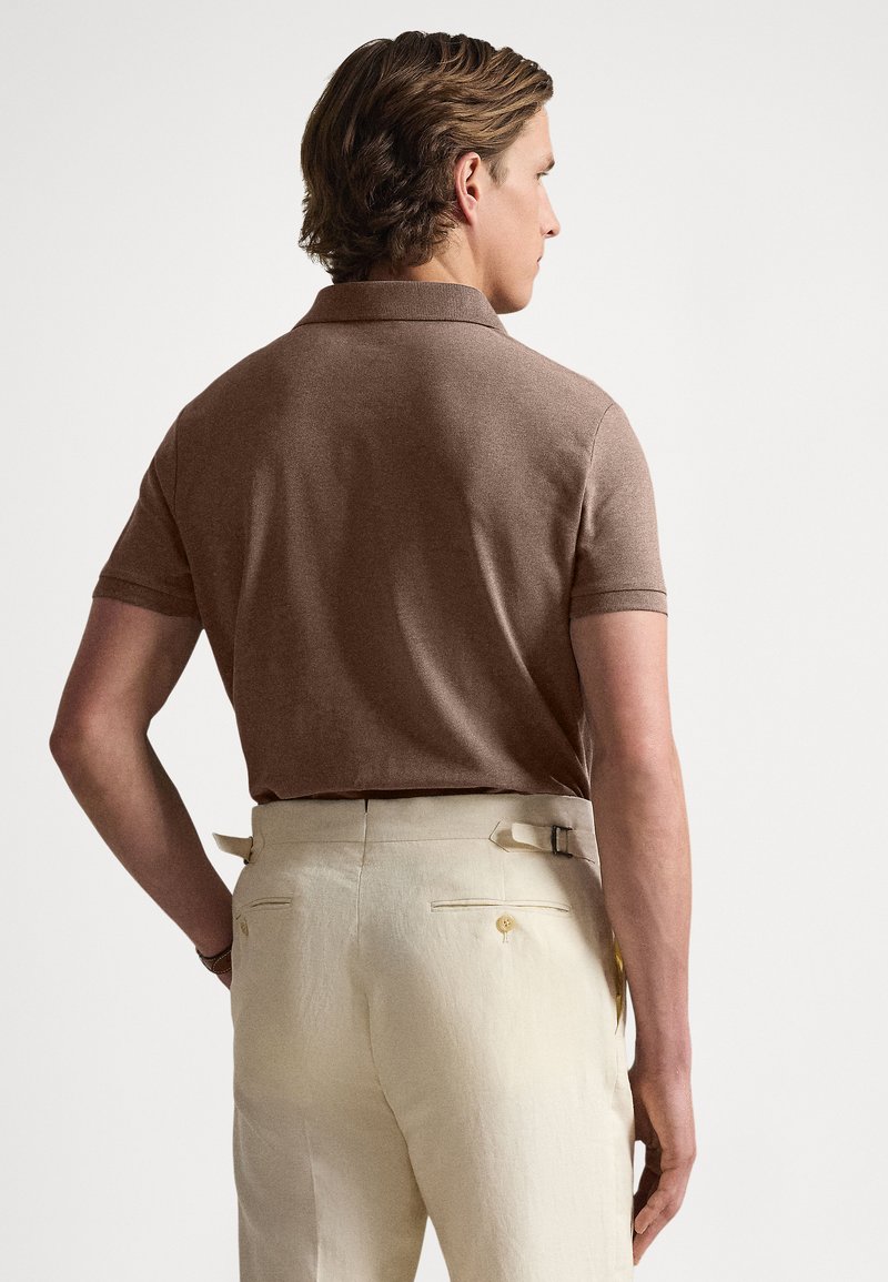 Braunes Poloshirt mit kurzen Ärmeln und einem gerippten Kragen, kombiniert mit hellen, maßgeschneiderten Hosen mit einer leicht geknöpften Gesäßtasche.