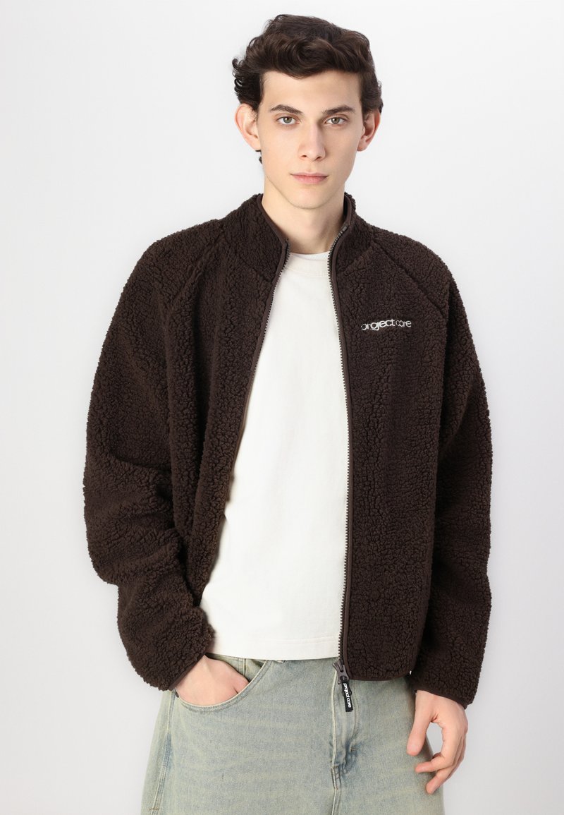 Brązowa polarowa kurtka z wysokim kołnierzem i zapinana na zamek błyskawiczny z przodu. Posiada logo na lewej piersi oraz miękką, teksturowaną powierzchnię.