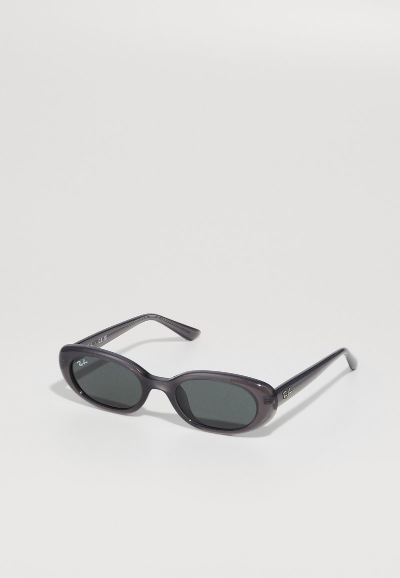 Juodos ovalios Ray-Ban saulės akinius su tamsiais objektyvais ant baltos fono, rodančius prekės ženklų logotipus ant objektyvo ir šventyklų.