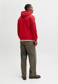 Sweat-shirt à capuche rouge avec des poignets côtelés, superposé à une chemise beige, assorti à un pantalon ample vert olive et des chaussures marron.