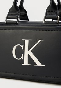 Μαύρη τσάντα χειρός από συνθετικό δέρμα με δομημένο σχεδιασμό, που διαθέτει λευκό ανάγλυφο λογότυπο 'cK' και ασημί μεταλλικά στοιχεία.