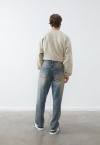 Hombre con pelo castaño, vestido con un sudadera beige, jeans azules descoloridos y zapatillas blancas, de pie frente a una pared blanca sobre un suelo gris.