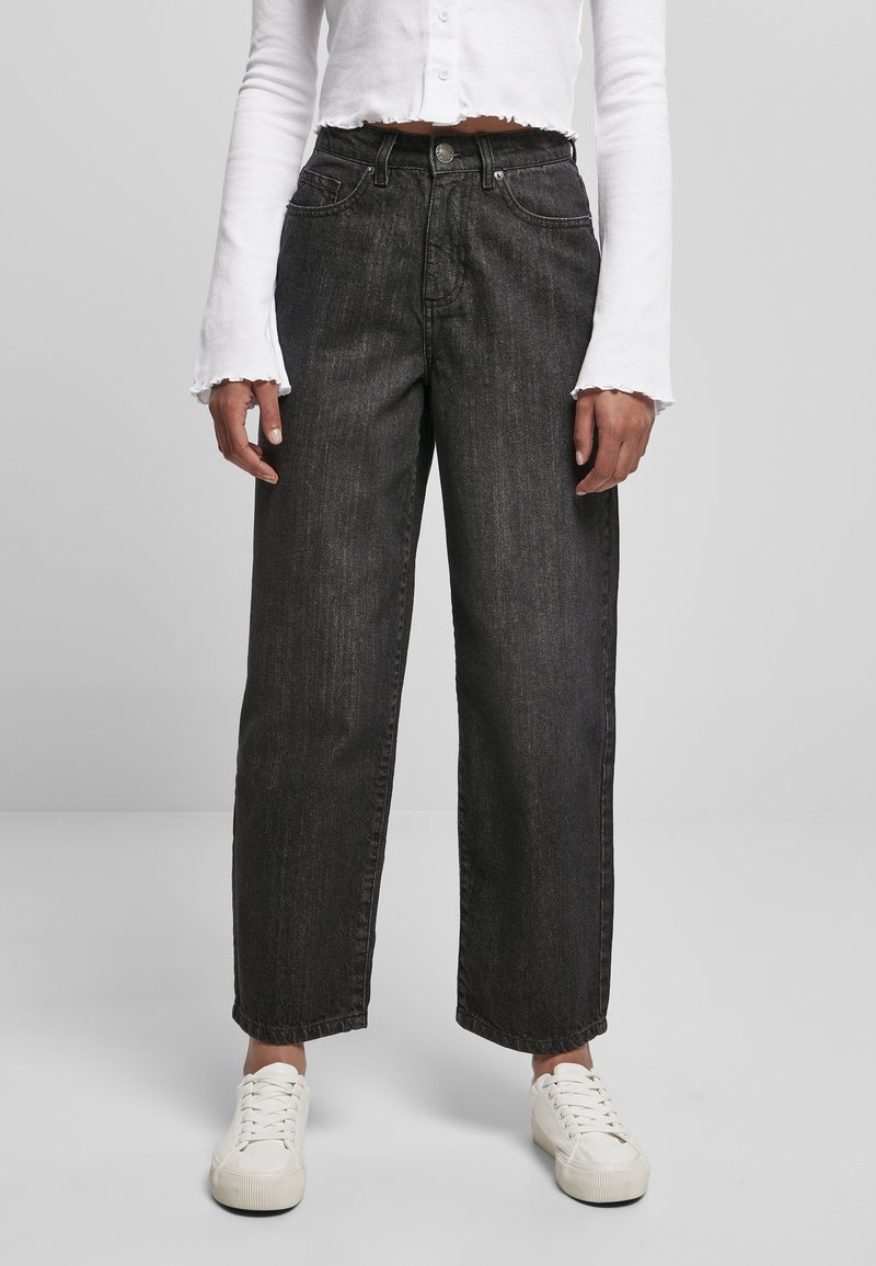 Personne portant un jean noir taille haute à coupe ample, des baskets blanches et un top court blanc à manches longues sur un fond uni.