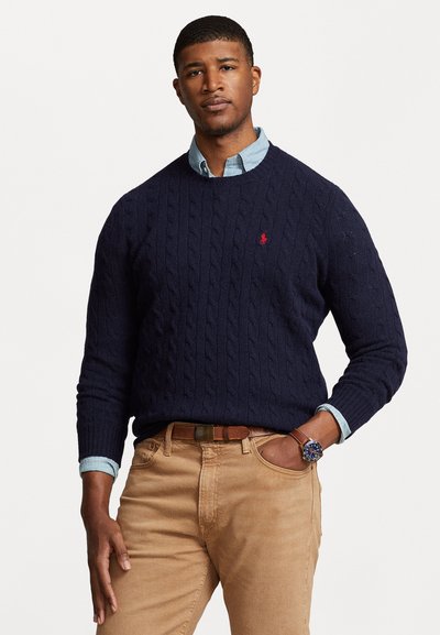 Marine kabelstrik sweater med rund hals, båret over en lyseblå skjorte. Tan bukser og et brunt bælte fuldender outfittet.