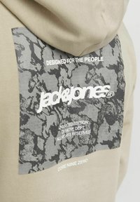 Beige hoodie featuring a textured dark gray graphic print on the back with the text "DESIGNED FOR THE PEOPLE" and brand details.