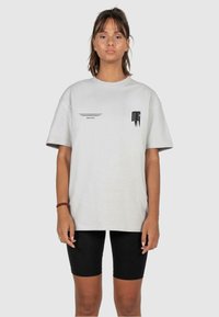 Helles graues Baumwoll-T-Shirt mit Rundhalsausschnitt, kurzen Ärmeln und schwarzen Text- sowie Logoakzenten auf der Vorderseite. Schwarze enganliegende Shorts.