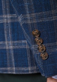 Niebieska marynarka w wzory z mankietami z teksturowanej tkaniny, z czterema brązowymi guzikami z logo oraz jasnym wzorem w kratkę.