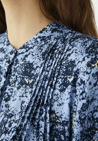 Modrá blůza s vzorem disponuje texturovaným materiálem, s záhyby na ramenou a knoflíkovým detailem u výstřihu, doplněná námořnickými a zlatými akcenty.