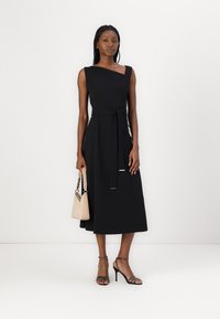 Robe noire sans manches avec un col diagonal et une taille ceinturée, présentant une silhouette fluide, associée à des talons noirs à lanières et un sac beige.