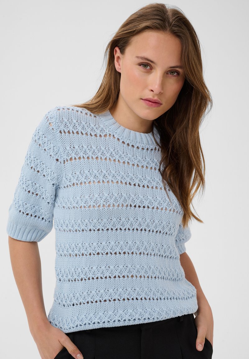 Jeune femme aux longs cheveux bruns portant un pull en maille bleu clair à manches courtes et un pantalon noir sur un fond uni.