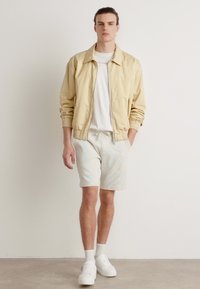 Giacca a zip beige chiaro, t-shirt bianca, pantaloni corti chiari e sneakers bianche; texture liscia, vestibilità casual e design minimalista.