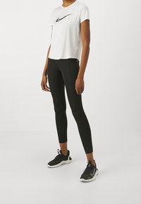 Bílý tričko s krátkým rukávem Nike s černým logem; v kombinaci s černými legínami a černými sportovními botami s bílou podrážkou.