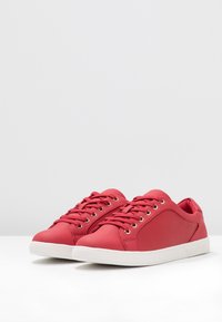 Chaussures de sport rouges en matière synthétique lisse avec un panneau latéral texturé, une semelle en caoutchouc blanche et des œillets métalliques pour les lacets.