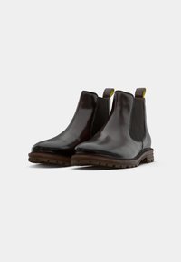 Botas Chelsea de cuero marrón con un diseño elegante, paneles laterales elásticos y tiradores amarillos en contraste. Suelas de goma con tacos para mayor agarre.