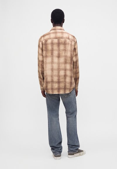 Homme debout, de dos, portant une chemise beige à carreaux à manches longues, un jean bleu et des baskets grises sur un fond uni.
