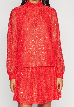 Rotes Spitzenkleid mit langen, puffigen Ärmeln, hohem, gerafftem Kragen und dezenten Paillettenverzierungen, getragen von einer Person mit geflochtenem Haar.