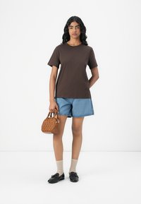 Hnědé bavlněné tričko, modré denimové šortky, béžová strukturální kabelka, béžové ponožky, černé mokasíny. Jednoduchý, neformální outfit uspořádaný na neutrálním pozadí.