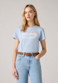 Šviesiai mėlynas medvilninis marškinėliai su baltu grafiniu "Levi's" logotipu. Suderinti su aukštu liemeniu, šviesiai mėlynomis džinsinėmis kelnėmis ir rudomis diržais.