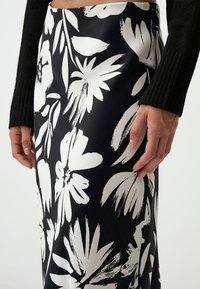 Zwarte rok met grote witte bloemenpatronen, met een gladde textuur en een strakke pasvorm, weergegeven naast een hand die op de stof rust.