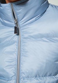 Light blue puffer jacket featuring a high collar, front zipper, and quilted texture with horizontal stitching. Silver zipper hardware detail.