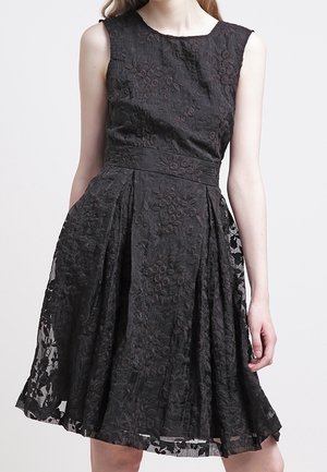 Vestido negro sin mangas, de largo hasta la rodilla, con superposición de encaje floral y cintura ajustada, llevado por una persona de pie frente a un fondo simple.