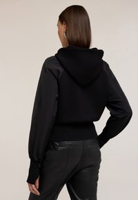 Felpa nera con cappuccio, realizzata in tessuto morbido, con maniche in satin e polsini a coste. Abbinata a pantaloni in pelle neri aderenti.