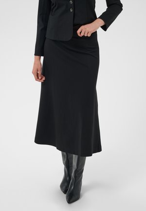 Femme portant une veste noire boutonnée, une longue jupe noire et des bottes hautes noires à bout pointu, debout avec une main près de la taille.