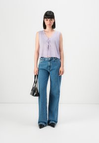 Kvinne med svart bob-klipp som har på seg en lavendel, ermløs strikket topp, blå vide jeans, svarte spisse sko, og holder en liten svart håndveske.