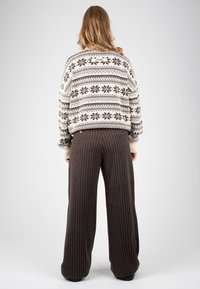 Personne aux cheveux longs portant un pull blanc et marron à motifs et un pantalon large marron foncé à rayures fines, debout devant un fond uni.