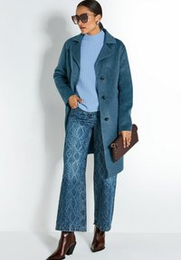 Vrouw in een teal wollen jas, lichtblauwe rib gebreide trui, met een gedrukt wijde broek van jeans, bruine leren laarzen, een geweven clutch vasthoudend, en oversized zonnebril.