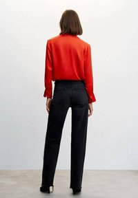 Camicia rossa di raso a maniche lunghe abbinata a jeans neri a vita alta, con gamba dritta e orlo grezzo. L'attenzione è rivolta alle trame e ai colori.