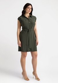 Abito camicia verde oliva con maniche corte, chiusura a bottoni sul davanti, due tasche sul petto e una cintura coordinata in vita. Tessuto dalla texture liscia.