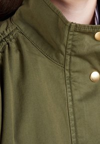 Olivgrön jacka med hög krage, prydd med gulddetaljer, broderade detaljer och en mjuk textur. Närbild på tyget och designens detaljer.