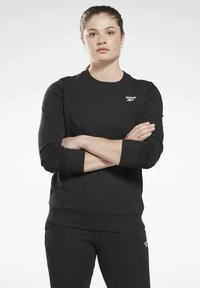 Crna dukserica s okruglim izrezom, rebrastim manžetama i rubom, s malim Reebok logotipom na lijevoj strani prsnog koša. Glatka tekstura i prilagođeni dizajn.