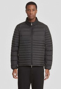 Zwarte pufferjack met horizontale gestikte patronen, hoge kraag, rits aan de voorkant en elastische mouwen. Draag het met zwarte broeken.