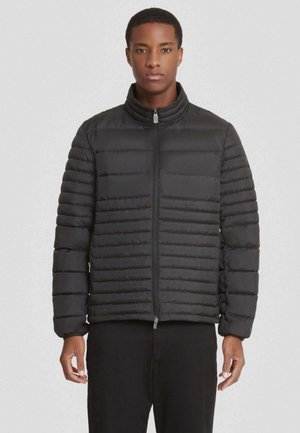 Giacca a vento nera con motivi a trapunta orizzontale, colletto alto, zip frontale e polsini elastici. Indossata con pantaloni neri.