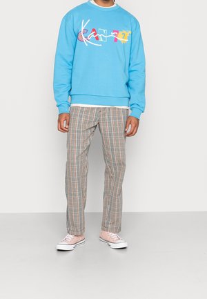 Felpa azzurra chiara con testo grafico multicolore "KANT?" e pantaloni a quadri beige e marroni con vestibilità rilassata, abbinati a sneaker chiare.