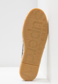 Suela negra de zapato con goma texturizada, con el logotipo "Superdry" en relieve y un color marrón claro. Forma redondeada con un perfil pronunciado.