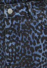 Tissu en denim bleu avec motif léopard noir, montrant un bouton en métal et une partie de la couture d'une poche sur un vêtement.