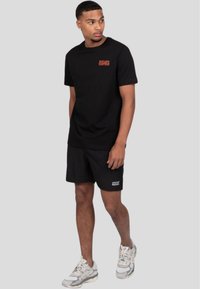 Sort bomulds t-shirt med rød tekst, parret med sorte shorts, hvide sko med grå accenter. Enkel design, afslappet pasform og minimal branding.
